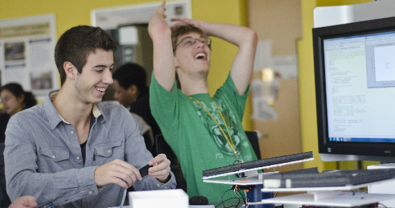 A student throws up his hands in joyful disbelief as another wields the controls of their engineering project.
